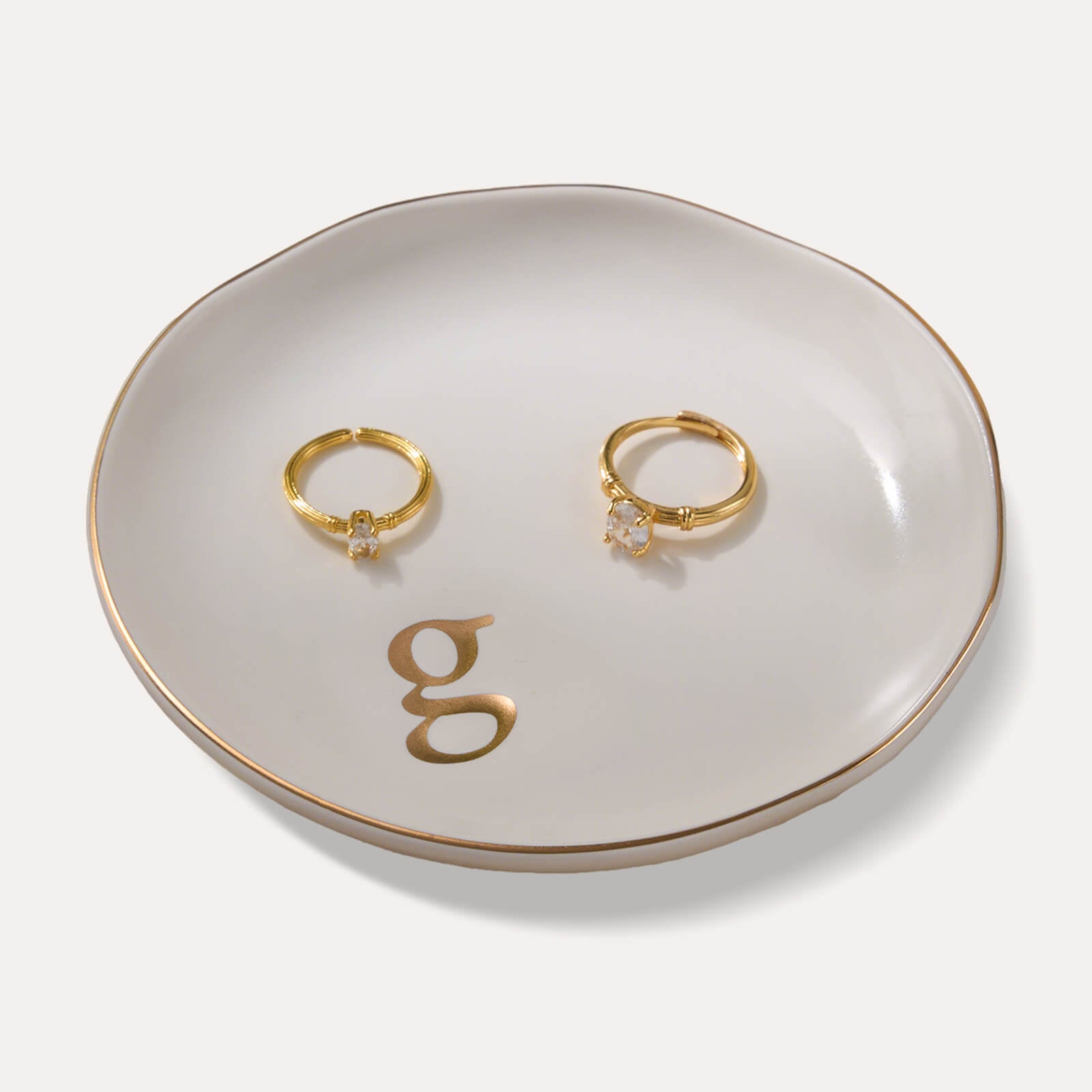 A Lolalet white ceramic jewelry dish featuring a gold "g" initial, elegantly holding an engagement ring, serving as a monogrammed ring holder.