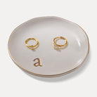 A Lolalet white ceramic jewelry dish featuring a gold "a" initial, elegantly holding an engagement ring, serving as a monogrammed ring holder.